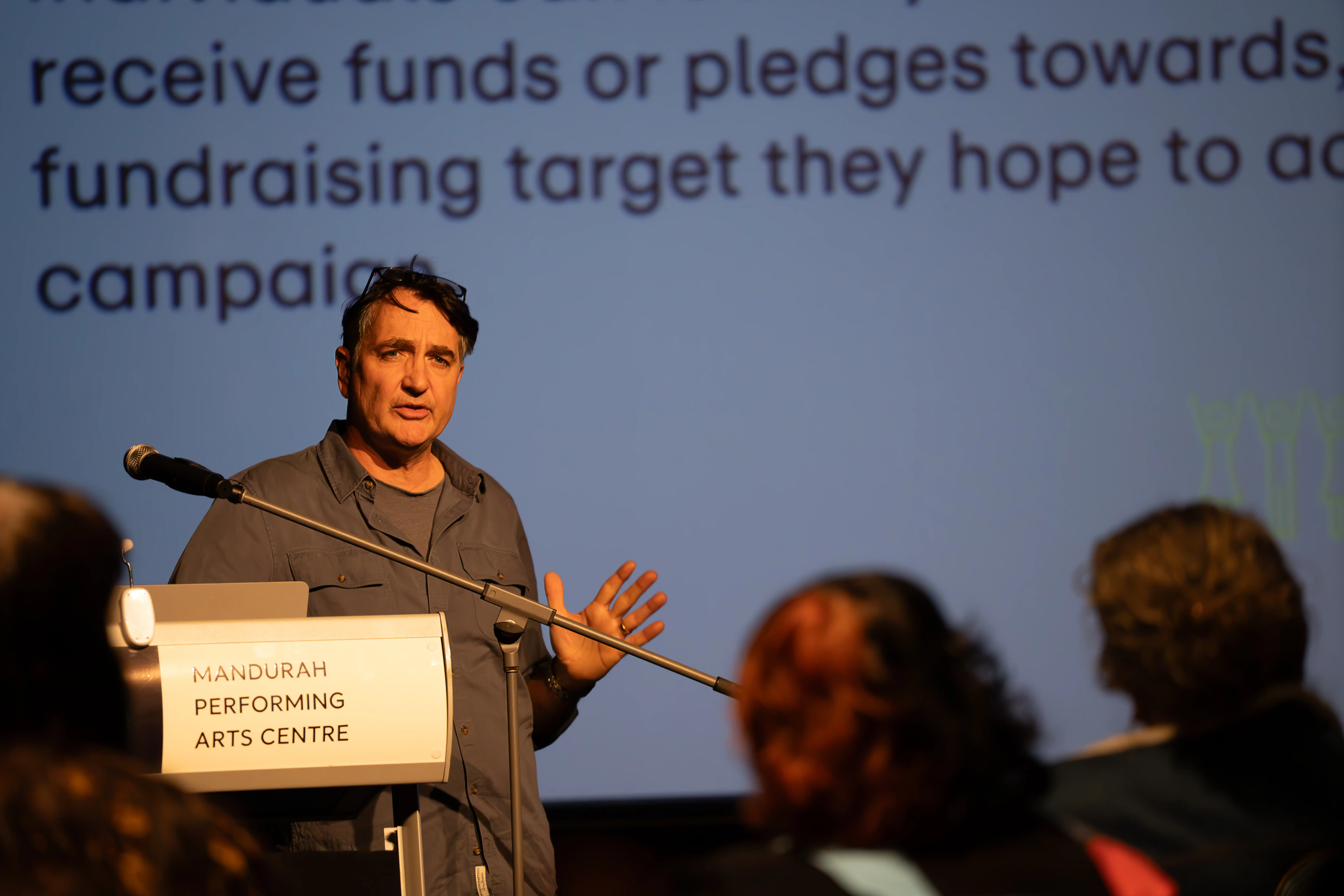 Presenter speaking at a podium in front of a projected screen while audience members listen.